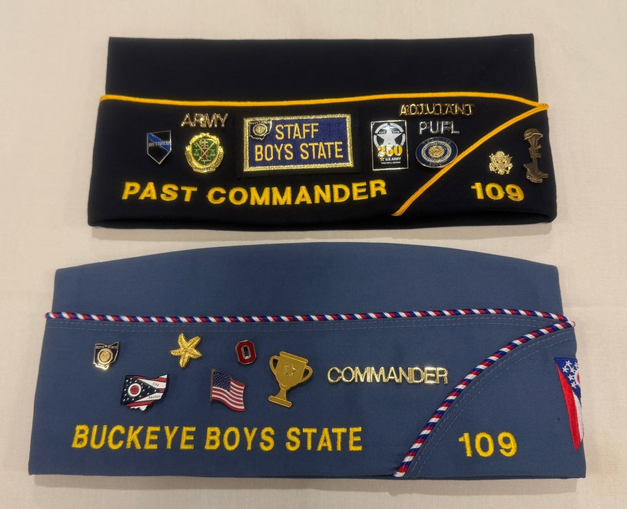 Two SAL Squadron garrison caps covered in pins, one reading 'Past Commander' and the other reading 'Commander' and 'Buckeye Boys State 109'